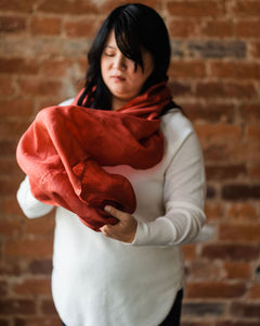 Asian woman wearing a cozy white sweater looking down and getting ready to loop russet red silk infinity scarf around her neck a second time.  Her face is slightly out of focus and her wavy black hair is caught under the section of scarf already along her neck.  Her bangs sweep low across her forehead