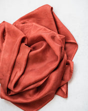 Top down view of russet red silk scarf gently piled in the middle of a white background, with gentle folds that create soft highlights and shadows.