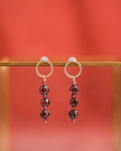 Delicate garnet earrings hanging from a slightly worn brass frame, against the russet red silk scarf in the background.  Only the earrings and the bar they're hanging from are in focus.  The earrings are gold filled open circle posts, each with a garnet drop.  The drops alternate four very small, faceted cube garnets with three larger and irregularly rounded, smooth garnets.