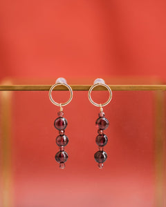 Delicate garnet earrings hanging from a slightly worn brass frame, against the russet red silk scarf in the background.  Only the earrings and the bar they're hanging from are in focus.  The earrings are gold filled open circle posts, each with a garnet drop.  The drops alternate four very small, faceted cube garnets with three larger and irregularly rounded, smooth garnets.