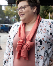 Plus size white woman with pixie cut medium brown hair, glasses, floral open front cardigan over a dusty pink shirt, looking off to the side and smiling with one hand on her hip.  She's wearing delicate garnet drop earrings and a 14x72" russet red silk scarf in a loop around her neck, with the ends tied into a poofy bow which rests along her bustline.