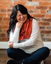 Asian woman wearing a cozy white sweater, dark jeans, black booties and russet red silk infinity scarf, sitting cross legged on wooden floor.  One elbow is propped on her knee with her arm bent up so she can lean her chin against her hand.  Her other arm crosses around so her hand can cup that elbow. Her wavy black hair falls down her back and over one shoulder, and her bangs sweeping low across her forehead.  She has a slender silver nose ring.  She's grinning.