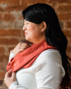 Profile shot of Asian woman wearing a cozy white sweater and arranging russet red silk infinity scarf around her neck, wavy black hair falling down her back and bangs sweeping low across her forehead.  She has a slender silver nose ring and delicate garnet drop earrings.