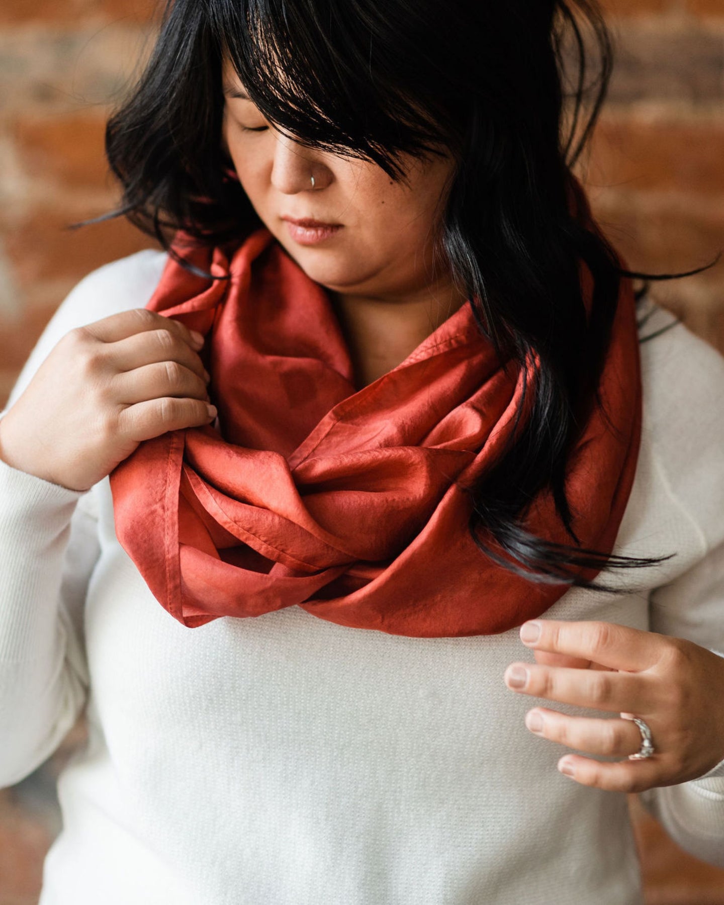 Asian woman wearing a cozy white sweater looking down and arranging russet red silk infinity scarf around her neck, wavy black hair falling over her shoulder and bangs sweeping low across her forehead