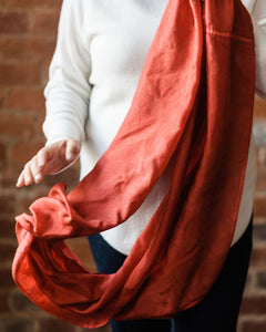 Torso shot of  woman holding russet red silk infinity scarf.  She's wearing a cozy white sweater and dark jeans.  She's holding one end of the infinity scarf up, almost out of frame, and her other hand is in the middle of the frame, slightly motion blurred as she drops the other end of the scarf.