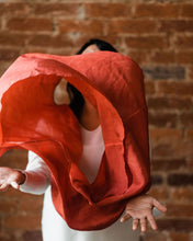 Woman tossing russet red silk infinity scarf.  She's wearing a cozy white sweater and her hands face up  at waist height to catch the scarf as it comes down.  The billowing scarf hides her face, but parts of her forehead, neck and wavy black hair peek around and through the loop of the infinity scarf.