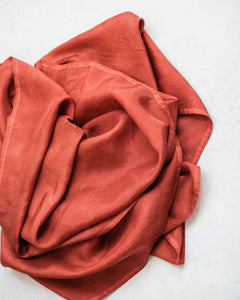 Top down view of russet red silk scarf gently piled in the middle of a white background, with gentle folds that create soft highlights and shadows.