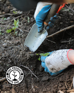 Person wearing blue and white gardening gloves, putting the tip of a hand trowel into dirt with one hand and holding a small seedling to the side, out of the way, with the other.  There is an overset black and white graphic with text in the lower left hand corner.  The words form a circle around a silhouette of a sapling with four leaves rising out of a mound of dirt. The words say "This product plants one tree". 
